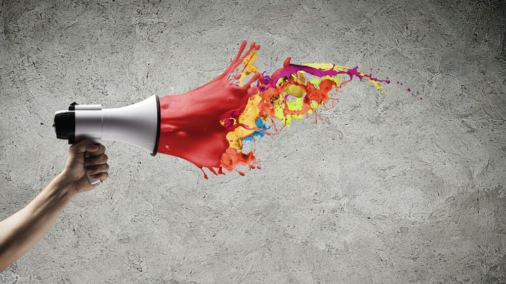 Hand holding a megaphone. The megaphone is white and red, and the red part fades off into multicolored paint splatters