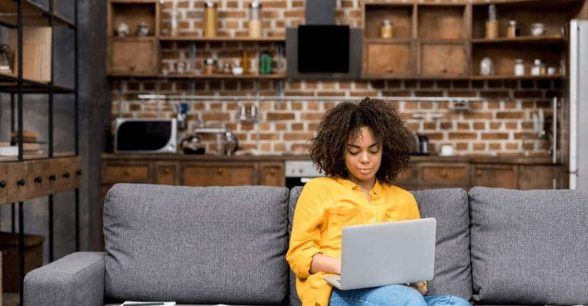 Young female-presenting person of color sitting on their couch with a laptop on their lap, working.