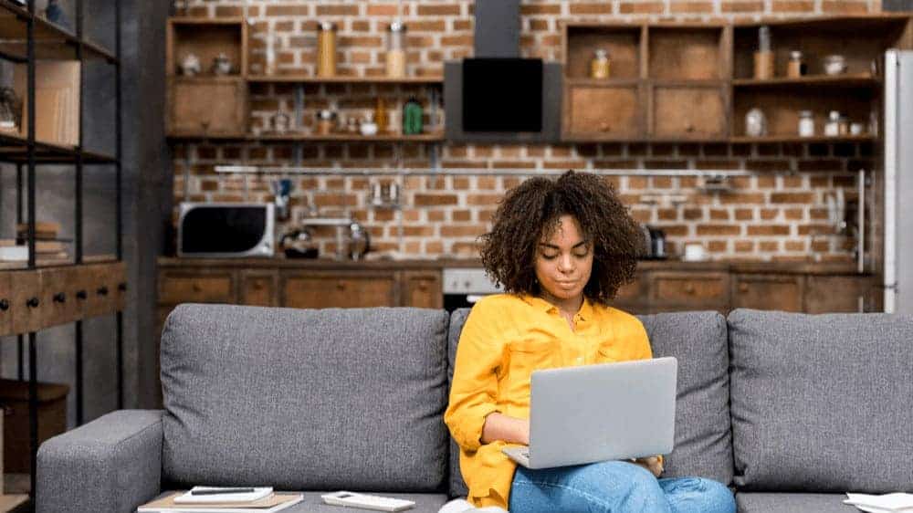 Young female-presenting person of color sitting on their couch with a laptop on their lap, working.