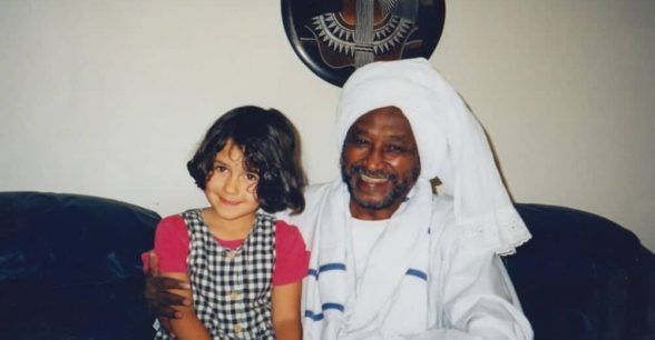 The author as a young child sitting on the lap of her Sudanese grandfather, who is dressed in a traditional white jellabiya and headwrap.