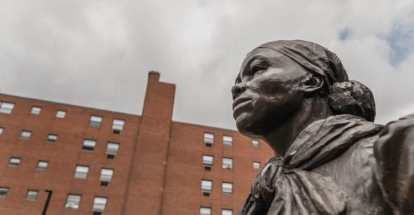 Against a background of a cloudy sky and brick building is the head of a statue of Harriet Tubman, taken from a lower angle so that the statue exudes strength and power.