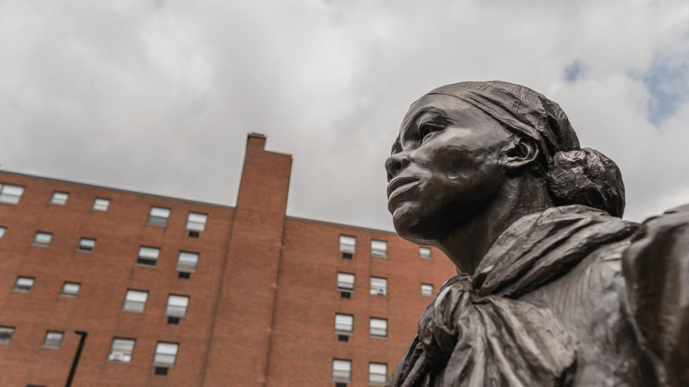 Against a background of a cloudy sky and brick building is the head of a statue of Harriet Tubman, taken from a lower angle so that the statue exudes strength and power.