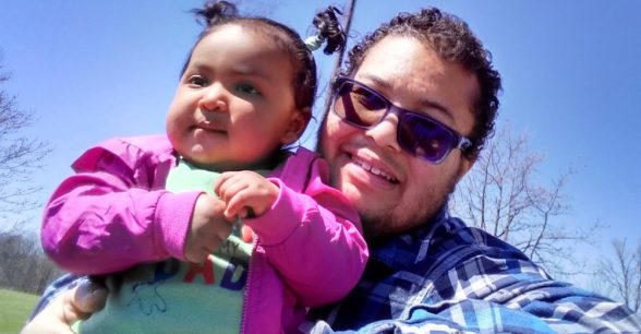 Photo of Kris and his daughter. Kris is a biracial man who has short black curly hair and tinted glasses. He is smiling while sitting holding his 11 month old daughter who has short black hair in two ponytails and is wearing a pink jacket on a sunny day at a park.