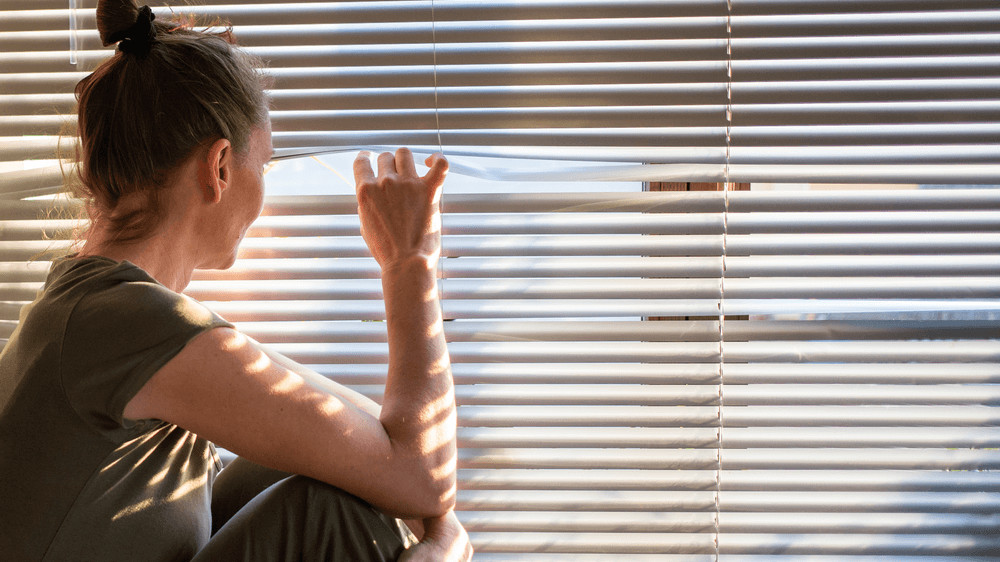 A white woman sitting next to closed blinds, using her fingers to open them and peek through the window to outside.