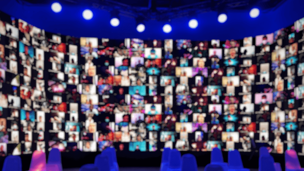 A wide rounded screen of blurred faces, all on webcam, under a row of spotlights.