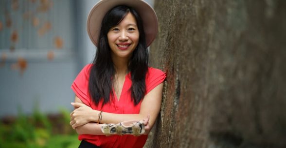 Photo of Tiffany, an Asian woman with dark hair below her shoulders, leaning against a tree. She is wearing a hat and a red top. Her arms are crossed, and she is wearing a splint.