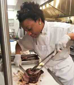 Photo of Hadassah, a black chef, in a restaurant kitchen, slicing a brisket.