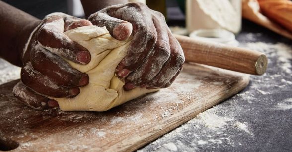 A Black person's hands kneading dough on a floured surface.