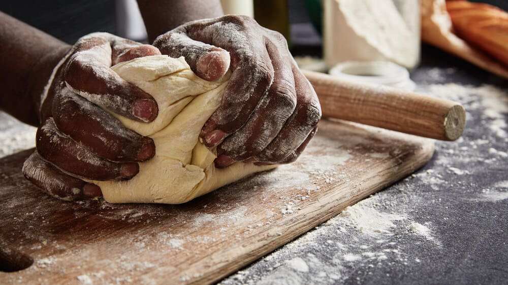 A Black person's hands kneading dough on a floured surface.