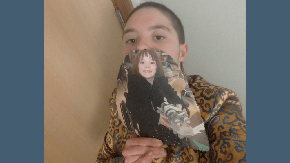 A young white woman with short hair, standing against a wall. In front of her face, she is holding a picture from when she was 8 years old, sitting in a wheelchair, smiling.