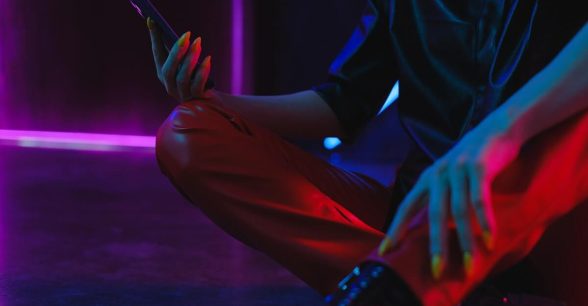 The bottom half of a person sitting on a floor, holding a cell phone in their hand. They're in a black shirt and red pants, and the lighting surrounding them is dark purple.