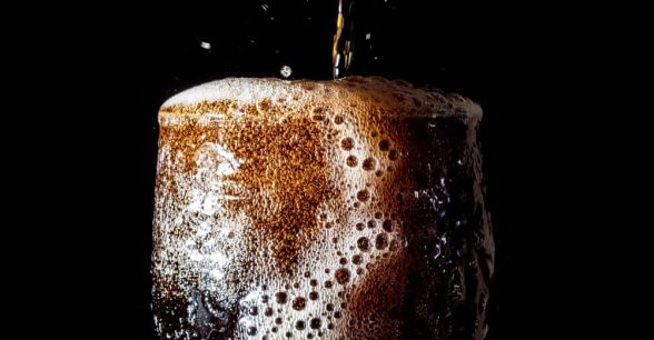 A clear glass overflowing with dark-colored, fizzy soda being poured from above. Plain black background.