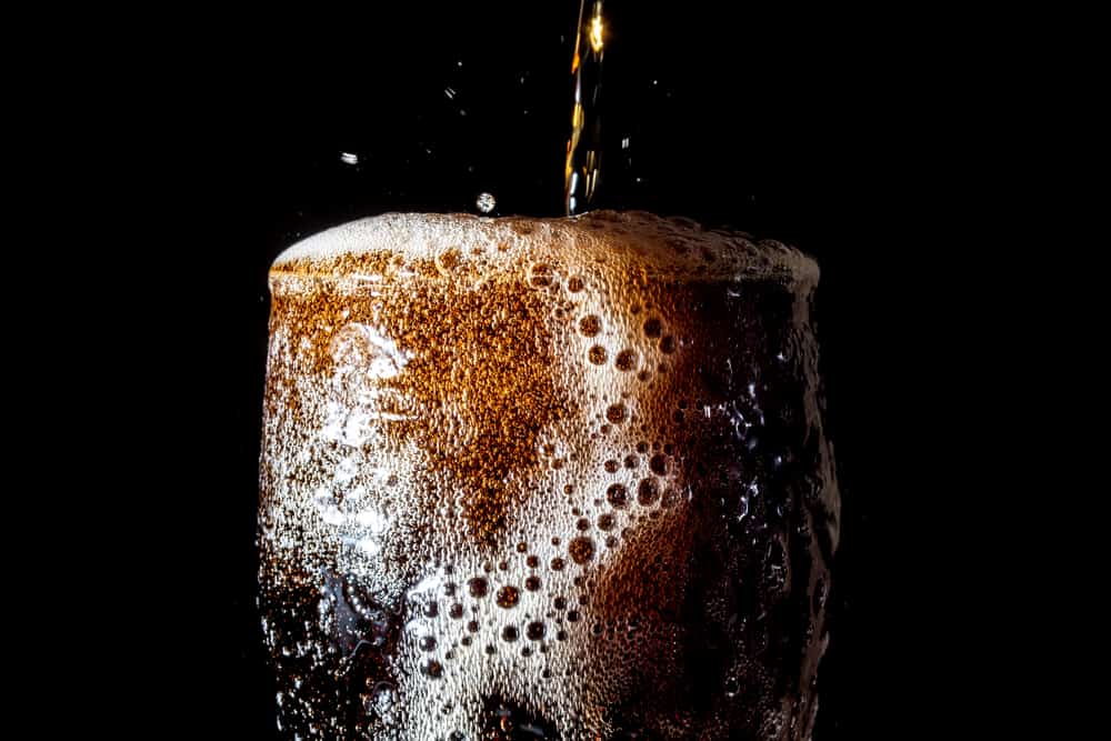 A clear glass overflowing with dark-colored, fizzy soda being poured from above. Plain black background.