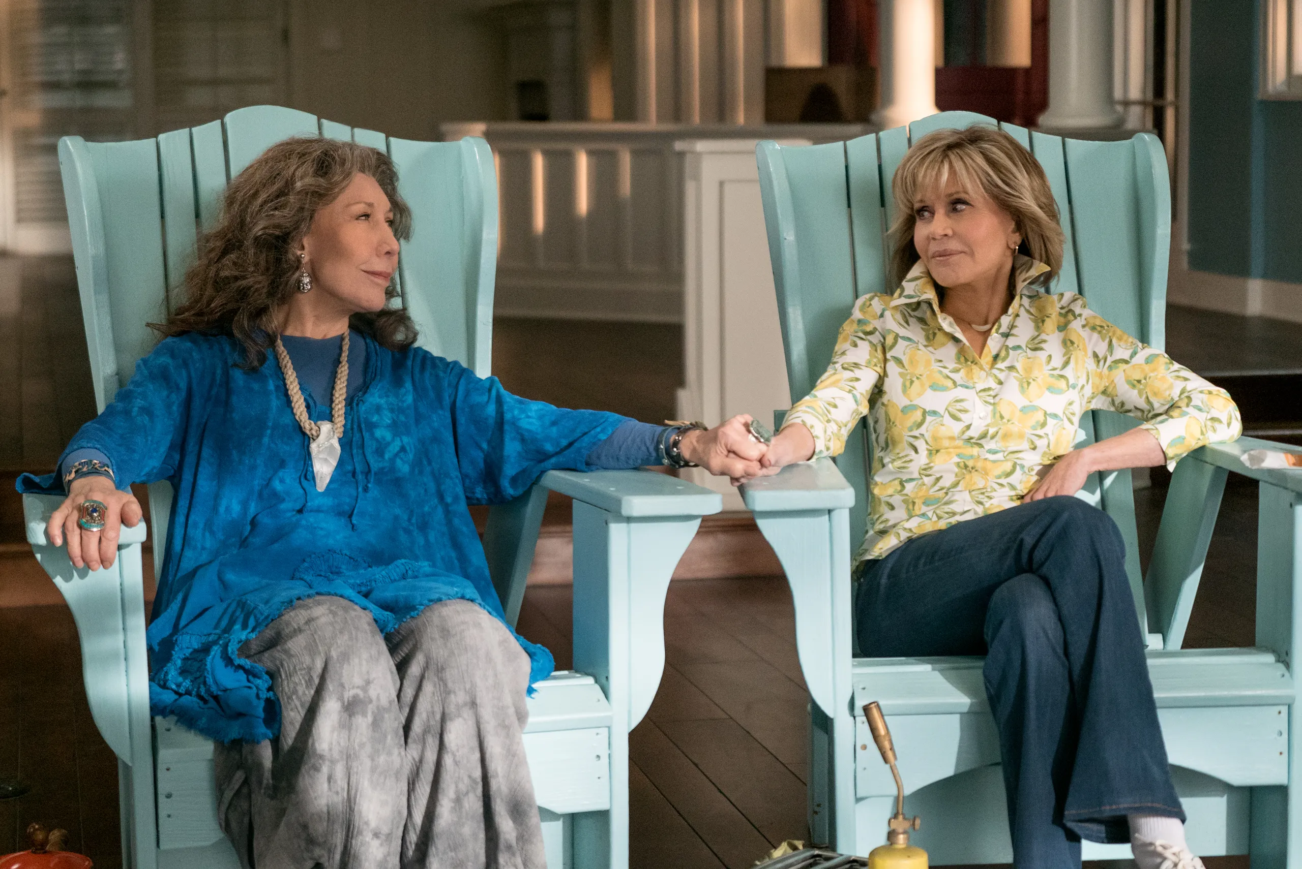 Two older white women, characters Grace and Frankie, sitting outdoors in separate patio chairs while holding hands and gazing at each other.