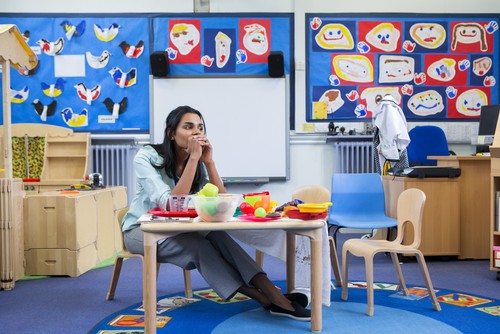 Stressed brown-skinned teacher with long, straight, dark hair in a colorful classroom, sitting at the toy table.