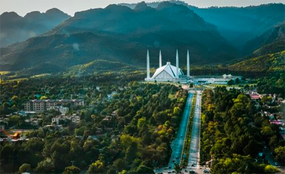 Daytime, an aerial view of Islamabad, Pakistan. It's mostly green tree tops with buildings and roads. Large mountains in the background.