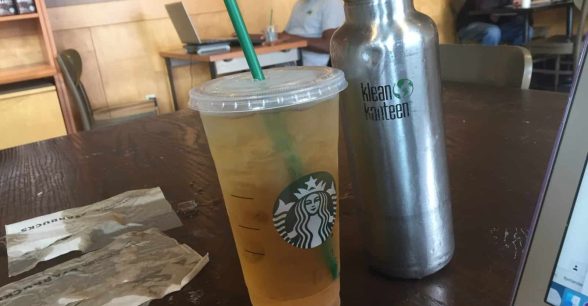 Indoors. A plastic Starbucks container of iced green tea and a dented, silver reusable Klean Kanteen brand water bottle sit on a dark wood grain table. To the left of them, two brown Starbucks napkins are soaked with liquid and ice. Other customers in the background.