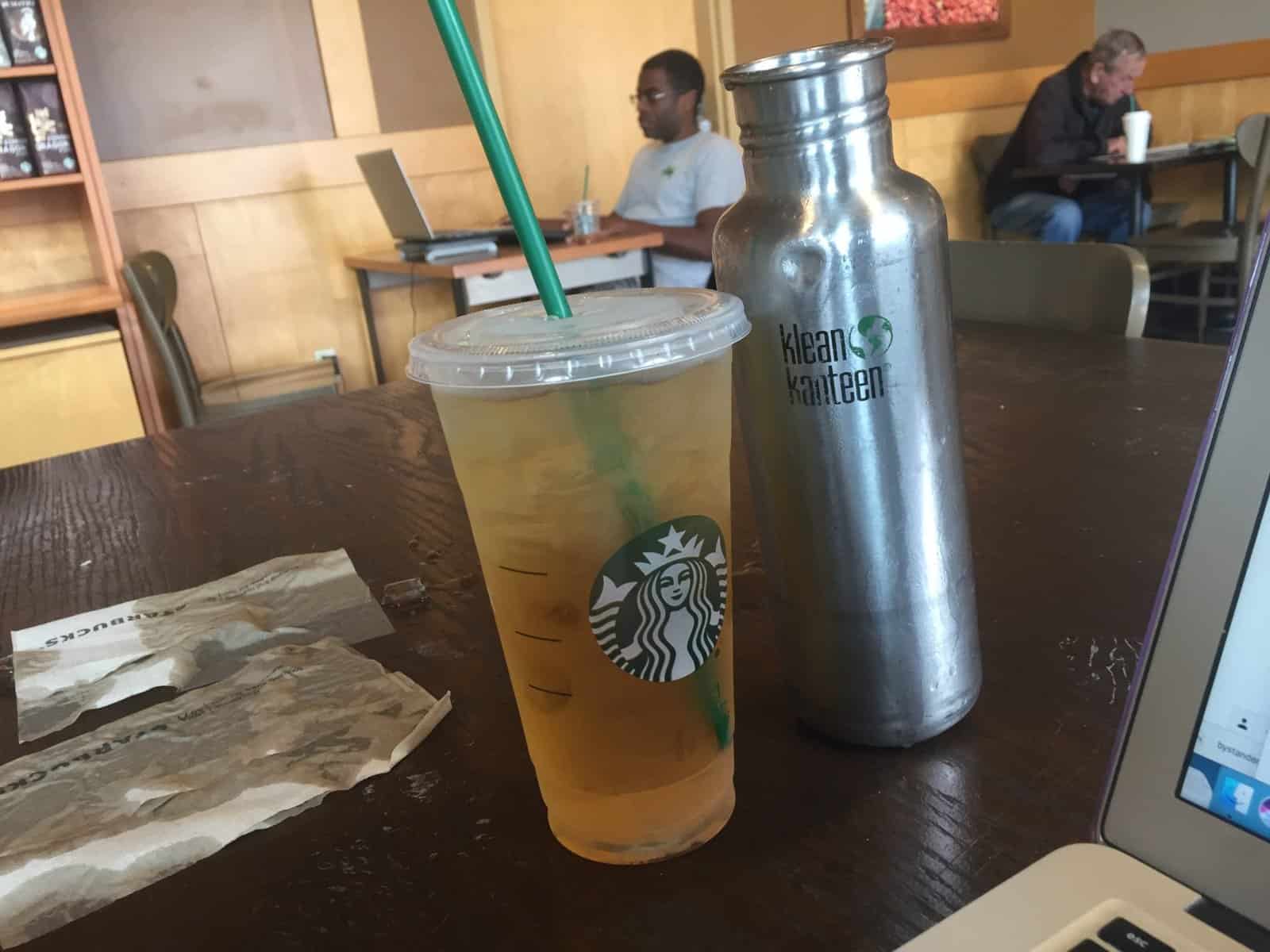 Indoors. A plastic Starbucks container of iced green tea and a dented, silver reusable Klean Kanteen brand water bottle sit on a dark wood grain table. To the left of them, two brown Starbucks napkins are soaked with liquid and ice. Other customers in the background.