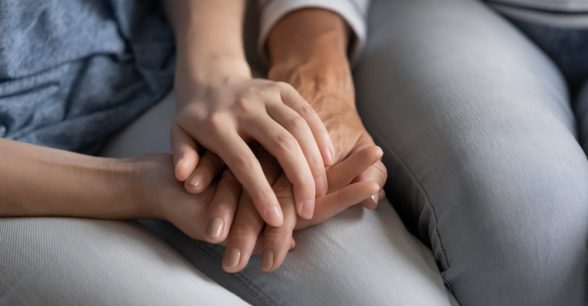 Close up of three hands. An elder's hand lays between the hands of a younger person's two hands. All three fair-skinned hands rest on the younger adult's lap.