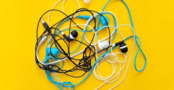 Against a bright yellow background, a small pile of earbud headphones, in different colors, is tangled up together.