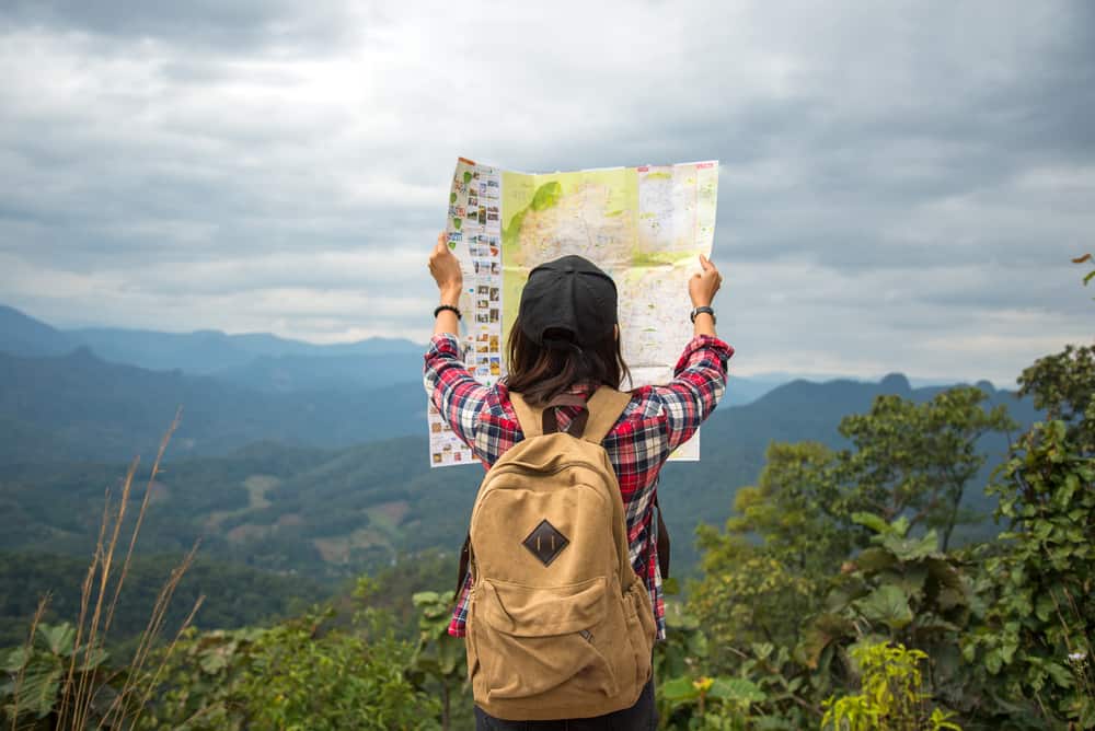 A dark-haired person stands at or near the top of a mountain range. Facing with their back to the camera, they're wearing a tan backpack as they hold up a very large map toward the sky to examine the map. They're surrounded by greenery and a cloudy sky.