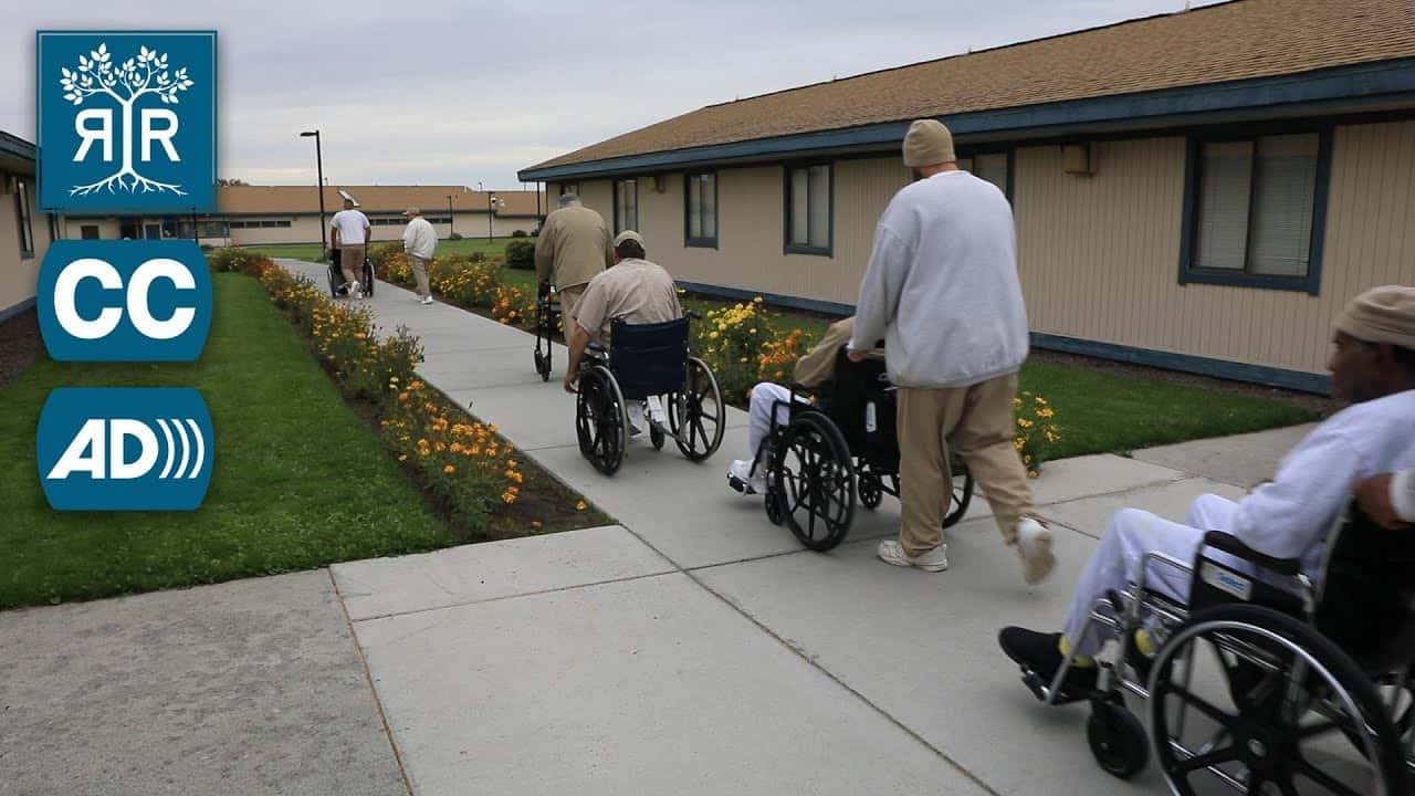 A row of inmates move down a walkway. Several are in wheelchairs. Another uses a walker.