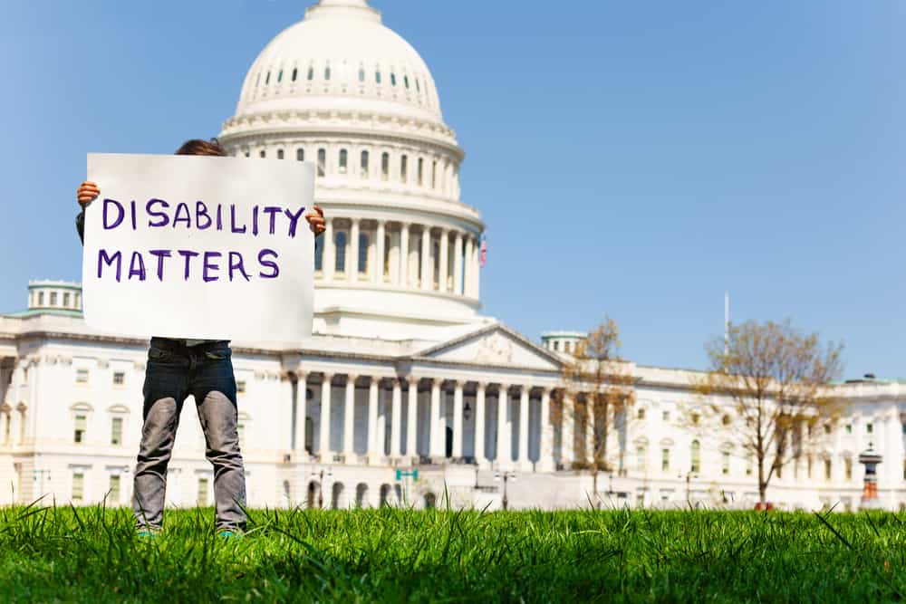 Outdoors, blue sky day. A person dressed in gray denim jeans stands in front of the U.S. Capitol building, holding up a sign that covers their face. It reads: "Disability matters."