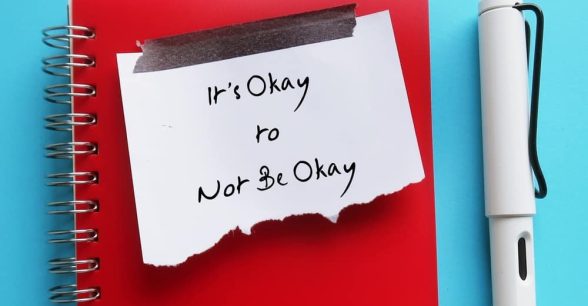 A red, spiral notebook and white pen against a light blue background. A white, torn off post-it note is taped to the notebook. It reads: "It's okay to not be okay."