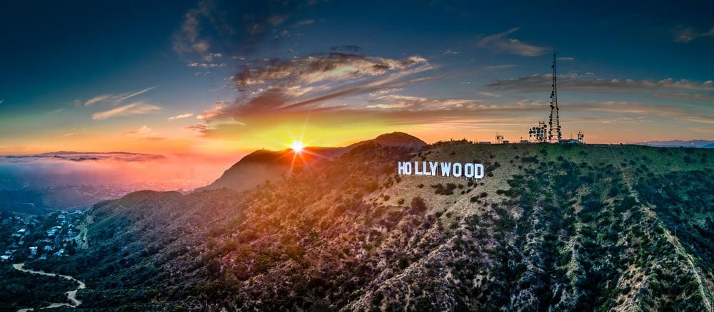The famous Hollywood sign in big, white block letters set on a mountainous, grassy terrain during either a sunrise or sunset.
