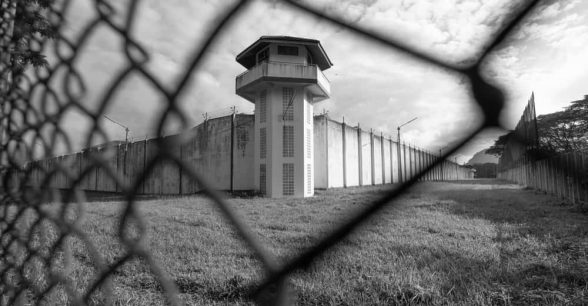 Outdoors, day. A prison watchtower separated by a wired fence. The camera is close up on the fence.