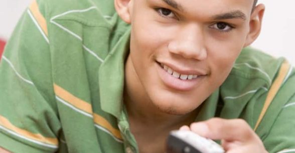 A light-skinned Black teenager in a green, striped, short-sleeved shirt is lying down on a bed, facing the camera and smiling. He has a TV remote in his hand and is pointing it at the camera.