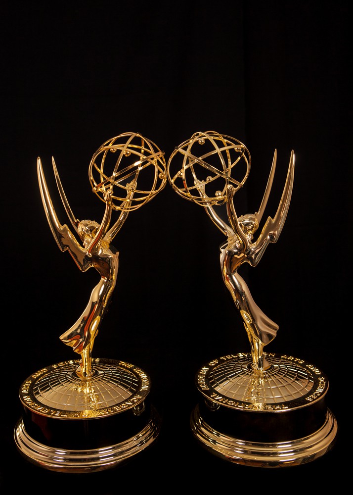 Black background. Two gold, winged Emmy statues stand facing each other.