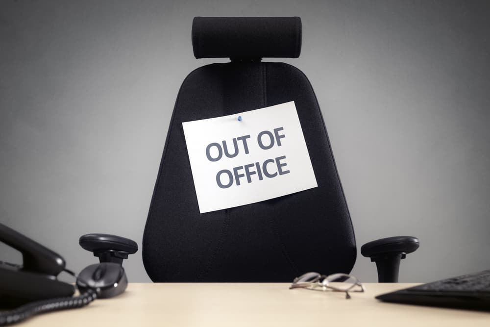 A comfortable-looking black office chair pulled up to a white desk with a black phone, black desktop keyboard, and a pair of glasses on it. A note attached to the chair has black text on white background that reads "out of office."
