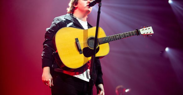Singer-songwriter-musician Lewis Capaldi, a white man in his 20s, wears a black jacket and pants and a white T-shirt as he stands in front of a microphone with an acoustic guitar wrapped around his neck, hanging across his chest. His eyes are closed and his lips are slightly parted as he sings into the mic.