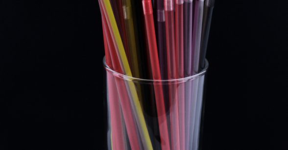 Black background. A clear drinking glass filled with multicolored plastic straws.