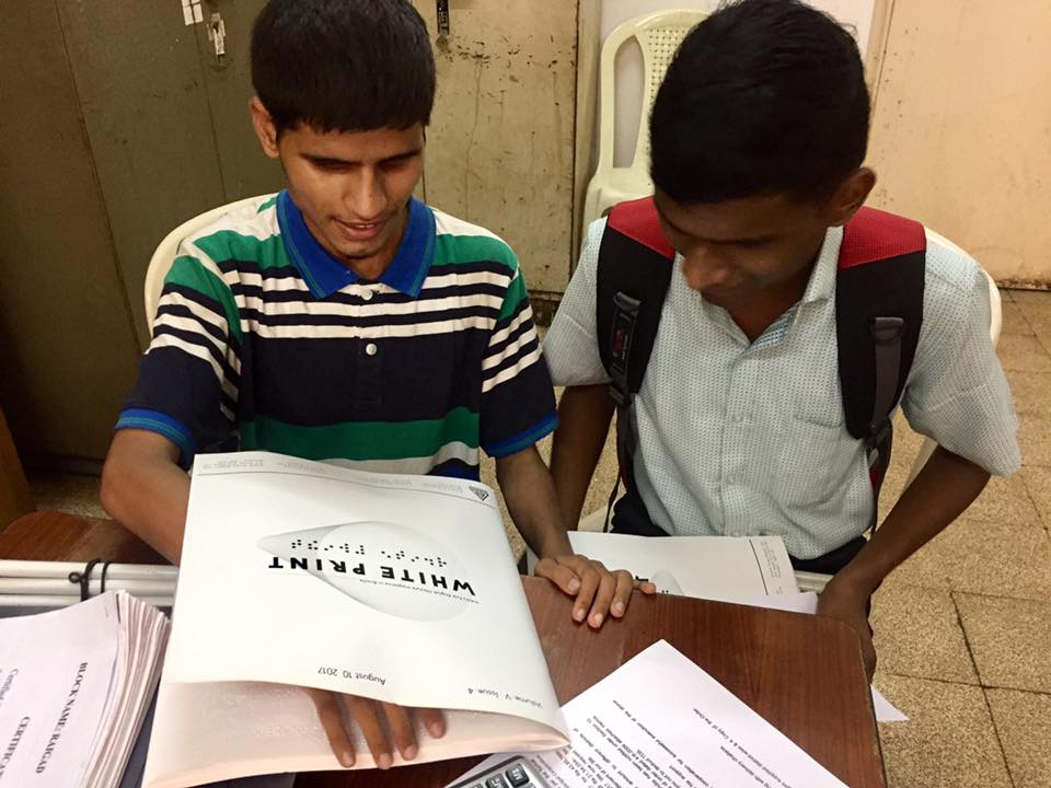 Indoors. Two dark brown-skinned Indian teens, one wearing a bookbag on their back, look down at copies of White Print mag that are located on a brown table.