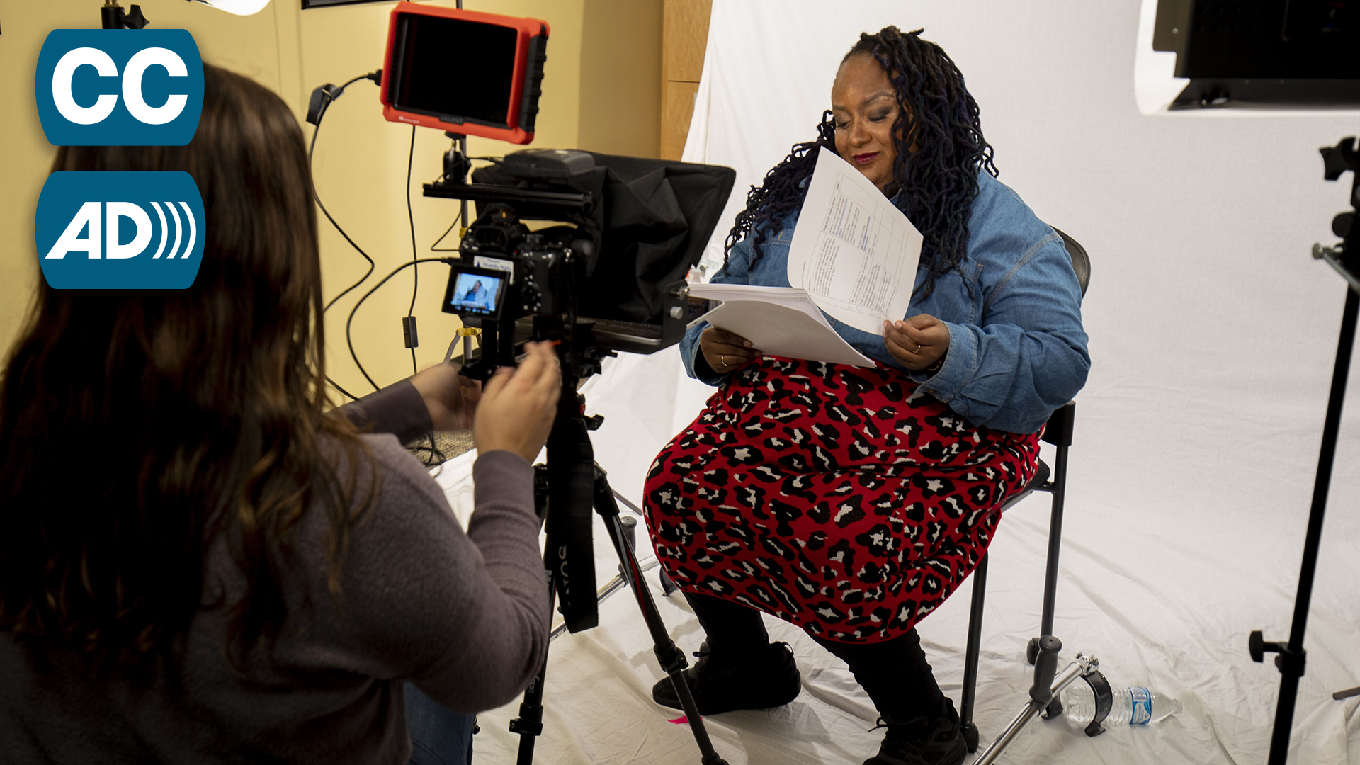 A young Black woman, Imani Barbarin, sits in front of a white backdrop reading a script. A camera, lights and a camera operator point toward Imani.