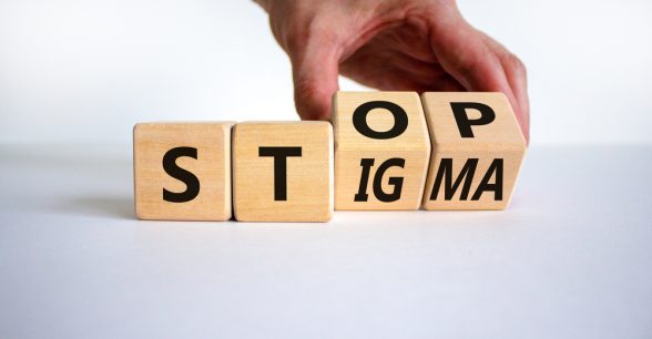 On a white background, a fair-skinned hand holds two of four wooden die. The die have letters on them that read, "Stop Stigma."