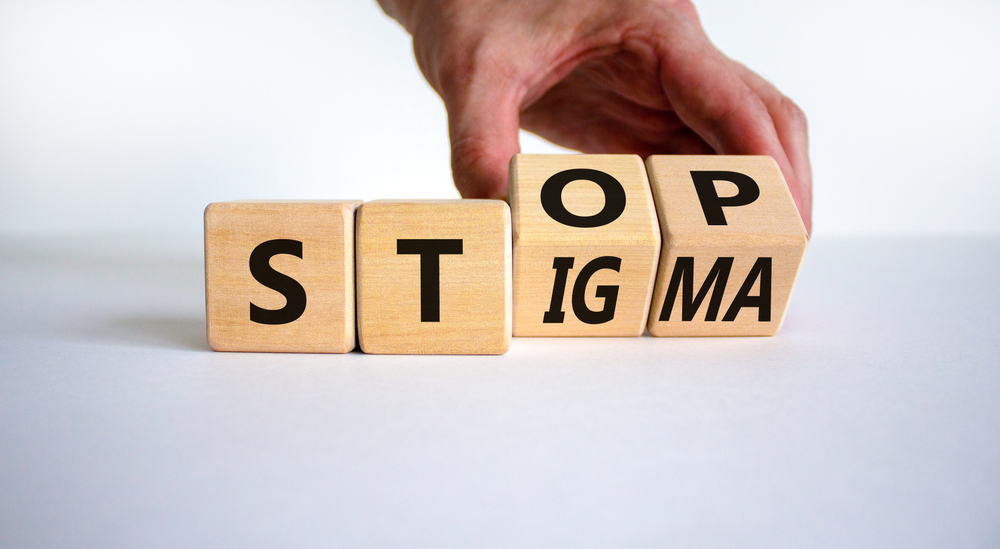 On a white background, a fair-skinned hand holds two of four wooden die. The die have letters on them that read, "Stop Stigma."