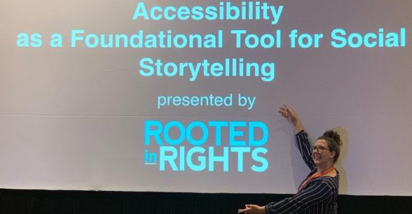 Allexa wearing a blue and white striped jumpsuit and bright yellow heels holds her arms up near a large screen that is projecting the words "Accessibility as a Foundational Tool for Social Storytelling presented by Rooted in Rights."