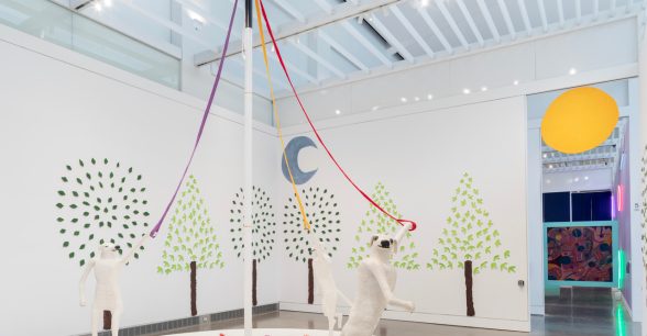 An installation containing three white, human-sized sculptures of dogs dancing on a circular platform and holding lavender, red, and orange dog leashes attached to a 15-foot tall white cane pole. The platform is covered in pink, magenta, and red papier-mâché flowers. The gallery walls are covered with trees made of light and dark green painted papier-mâché leaves. Hanging on the wall above the trees is a grey-blue crescent moon, and a large orange sun floating above the back entrance.