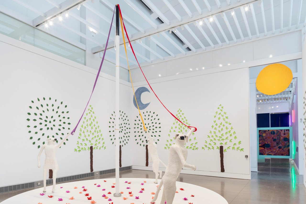 An installation containing three white, human-sized sculptures of dogs dancing on a circular platform and holding lavender, red, and orange dog leashes attached to a 15-foot tall white cane pole. The platform is covered in pink, magenta, and red papier-mâché flowers. The gallery walls are covered with trees made of light and dark green painted papier-mâché leaves. Hanging on the wall above the trees is a grey-blue crescent moon, and a large orange sun floating above the back entrance.